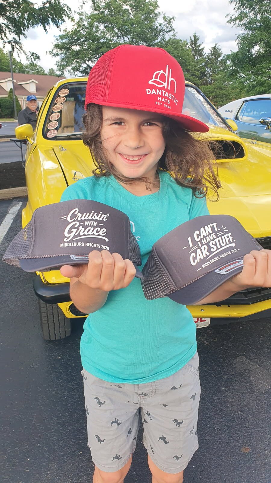 Daniel in a red Dantastic Hats cap holding two custom car-show hats in front of a yellow classic car.