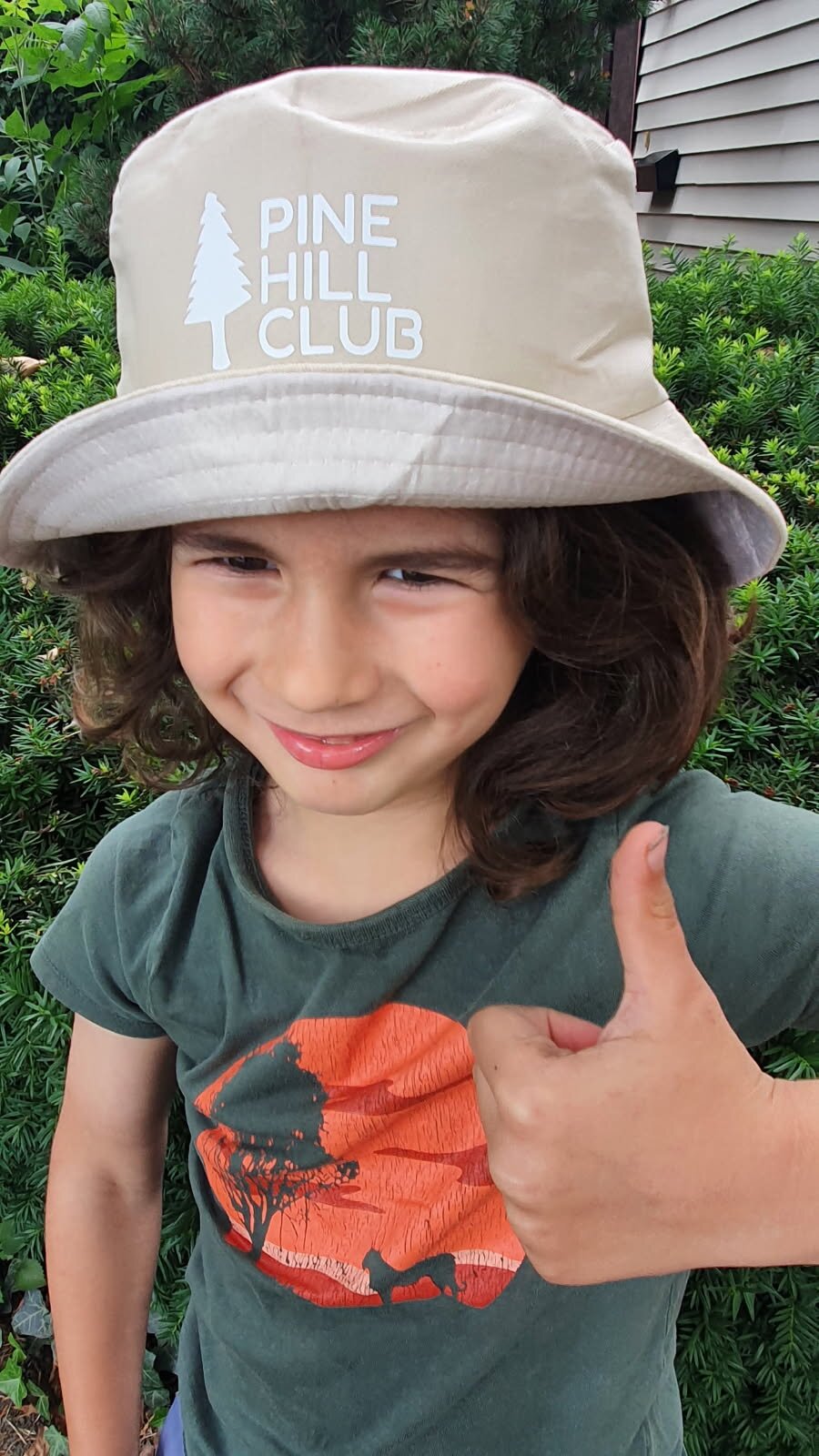 Daniel giving a thumbs-up in a tan Dantastic Hats bucket hat printed with a white 'Pine Hill Club' pine-tree logo.