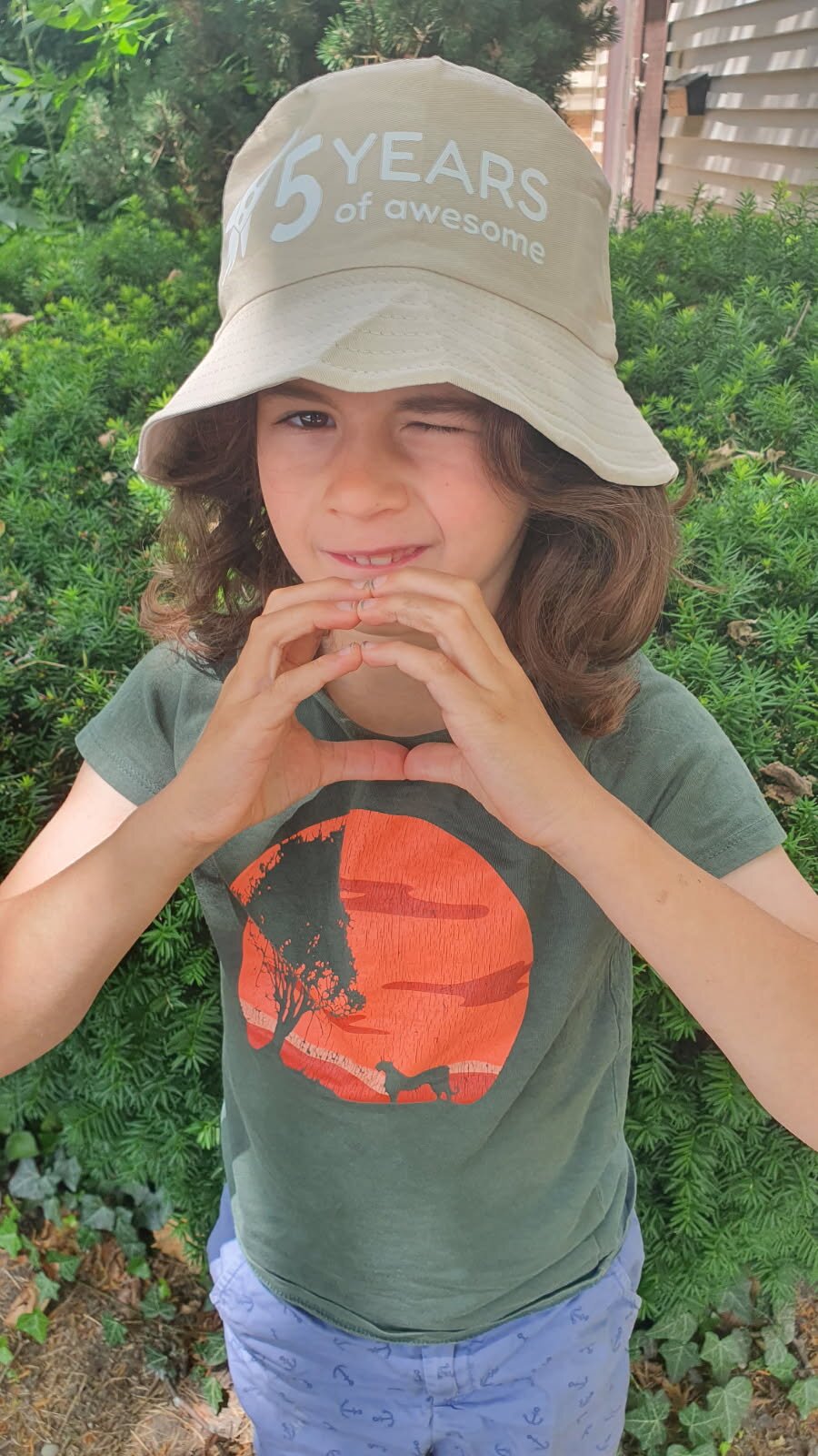 Daniel winking and making a heart with both hands in a tan Dantastic Hats bucket hat reading '5 Years of Awesome'.